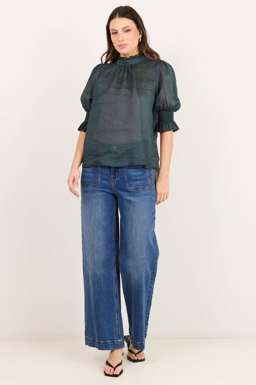 A woman modeling a dark green sheer puff-sleeve blouse with a gathered high neck, paired with wide-leg blue jeans and black sandals, standing against a plain off-white studio background.