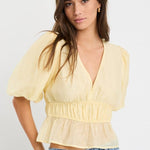 Woman wearing a butter yellow puff sleeve V-neck top with elastic shirring at the waist and a peplum hem, styled with light blue jeans.