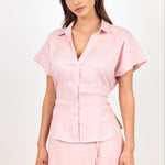 A woman modeling a light pink two-piece linen set consisting of a short-sleeve button-up shirt and a matching mini skirt, shot against a plain white studio background.