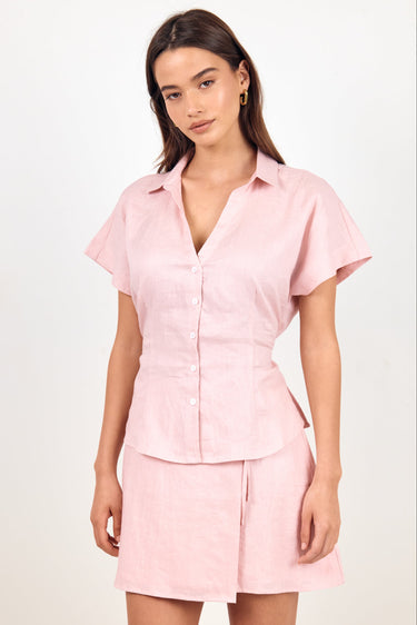 A woman modeling a light pink two-piece linen set consisting of a short-sleeve button-up shirt and a matching mini skirt, shot against a plain white studio background.