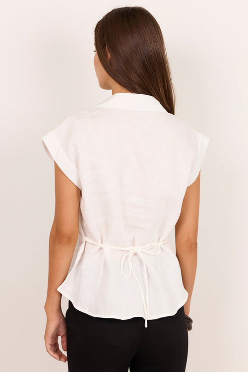 A woman viewed from the back wearing a white short-sleeve collared blouse with a tie detail at the waist and black pants, against a plain off-white background.