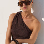 A woman wearing dark brown sunglasses, a dark brown textured one-shoulder top, and white cropped pants sits against a white wall, casting a strong shadow.