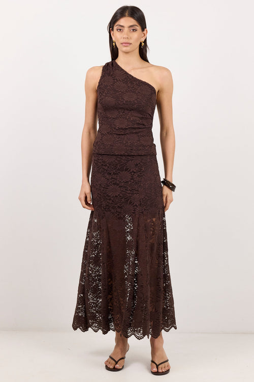 A woman modeling a dark brown one-shoulder lace maxi dress with a fitted bodice and flared skirt, paired with black strappy sandals, against a plain white studio background.