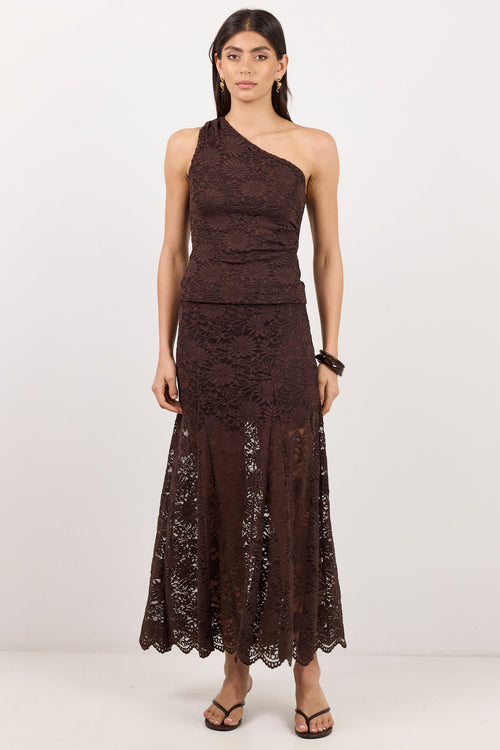 A woman modeling a dark brown one-shoulder lace maxi dress with a fitted bodice and flared skirt, paired with black strappy sandals, against a plain white studio background.