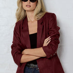 Person wearing a burgundy blazer and sunglasses against a white brick wall