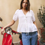 Woman in a white blouse and blue jeans standing next to a red scooter against a beige wall.