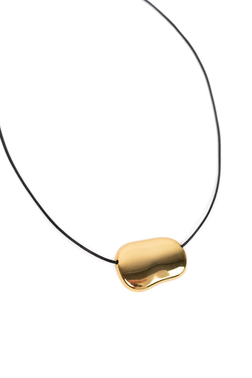 A minimalist gold pendant necklace with an organic, fluid bean-like shape on a thin black cord, shown against a solid white background.