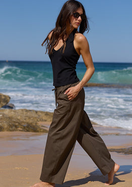 A woman walks barefoot on a sandy beach with ocean waves and rocks in the background, wearing a black sleeveless top and loose dark olive wide-leg pants.