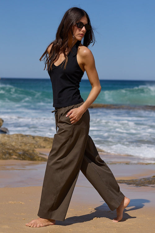A woman walks barefoot on a sandy beach with ocean waves and rocks in the background, wearing a black sleeveless top and loose dark olive wide-leg pants.