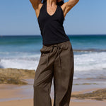 A woman stands on a sandy beach with the ocean and blue sky behind her, wearing a black sleeveless top and loose dark brown wide-leg pants.