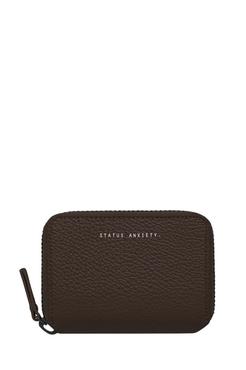 A dark brown pebbled leather zip-around wallet with 'STATUS ANXIETY' printed in white text on the front, shown on a plain white background.