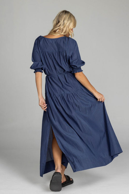 Woman wears a navy maxi dress with puff sleeves, a V-neckline, button front, waist tie, and side splits. The dress flows softly to the floor with a relaxed, feminine silhouette.