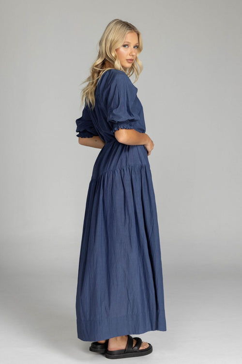 Woman wears a navy maxi dress with puff sleeves, a V-neckline, button front, waist tie, and side splits. The dress flows softly to the floor with a relaxed, feminine silhouette.