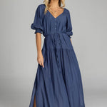 Woman wears a navy maxi dress with puff sleeves, a V-neckline, button front, waist tie, and side splits. The dress flows softly to the floor with a relaxed, feminine silhouette.