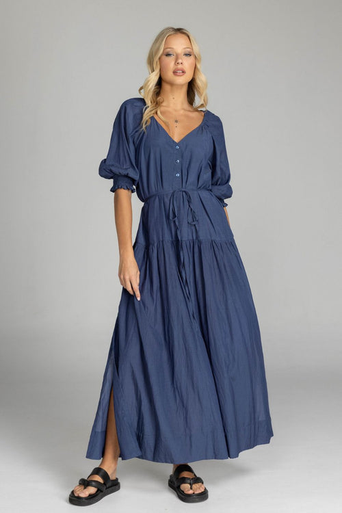 Woman wears a navy maxi dress with puff sleeves, a V-neckline, button front, waist tie, and side splits. The dress flows softly to the floor with a relaxed, feminine silhouette.