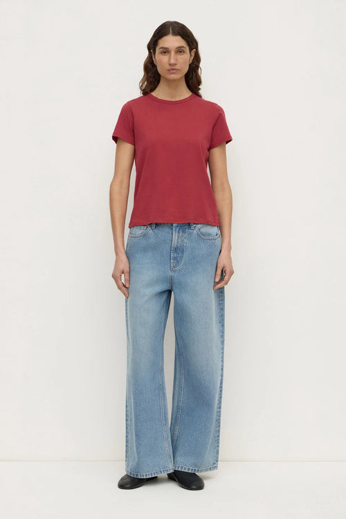 A woman stands against a plain white studio background wearing a short-sleeve crew-neck t-shirt in a muted brick red color, paired with light-wash wide-leg blue jeans and black shoes.