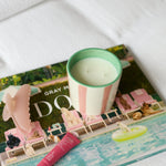 A styled product shot featuring a gray and pink striped candle with a mint green rim, resting on top of a colorful magazine on a white textured fabric surface.