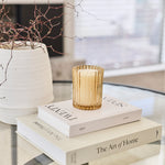 A gold-toned ribbed glass candle holder containing a white candle, styled on top of two white books on a glass table in a bright, modern interior.