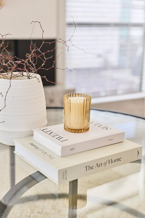 A gold-toned ribbed glass candle holder containing a white candle, styled on top of two white books on a glass table in a bright, modern interior.
