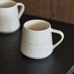 Two matte white ceramic mugs with gold lettering, one in the foreground and one blurred in the background, resting on a dark tabletop against a wooden wall.