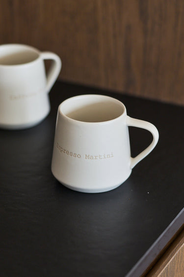 Two matte white ceramic mugs with gold lettering, one in the foreground and one blurred in the background, resting on a dark tabletop against a wooden wall.