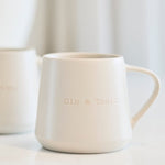 Two small white ceramic mugs with gold lettering on a light marble countertop, with a blurred white vase in the background.
