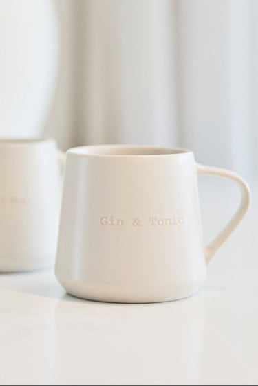 Two small white ceramic mugs with gold lettering on a light marble countertop, with a blurred white vase in the background.