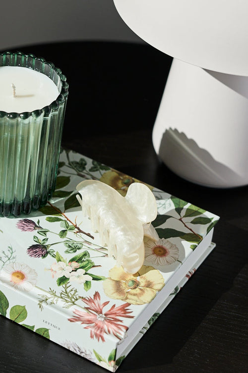 A styled interior scene showing a small round black table with a floral tissue box, a green ribbed candle holder, and a white mushroom-shaped lamp against a dark gray wall.