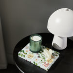 A styled interior scene showing a small round black table with a floral tissue box, a green ribbed candle holder, and a white mushroom-shaped lamp against a dark gray wall.