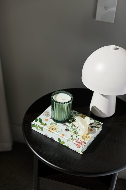 A styled interior scene showing a small round black table with a floral tissue box, a green ribbed candle holder, and a white mushroom-shaped lamp against a dark gray wall.