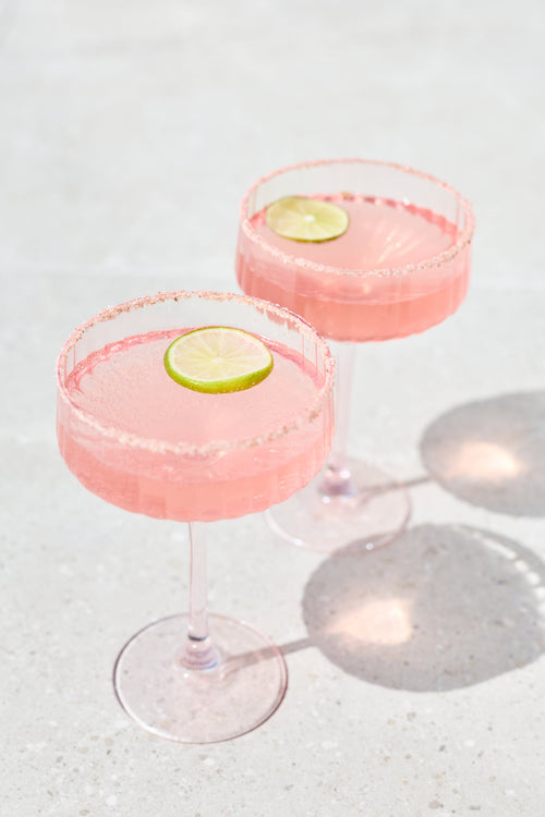 Two pink margarita cocktails in stemmed coupe glasses with salted rims and lime slices