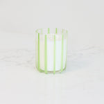 A white candle in a clear glass holder with light green vertical stripes, sitting on a white marble countertop against a white wall.