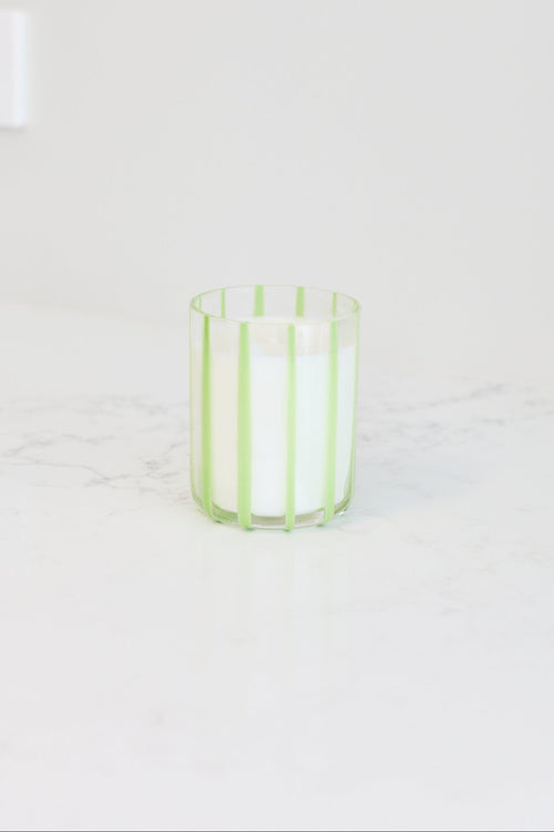 A white candle in a clear glass holder with light green vertical stripes, sitting on a white marble countertop against a white wall.
