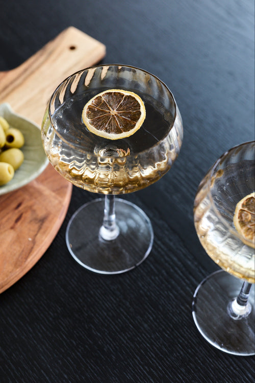Two clear stemmed coupe-style glasses on a dark tabletop, each containing a clear golden drink and topped with a dried lemon slice. A wooden board with green olives is partially visible on the left.