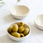 Three small off-white ceramic bowls arranged on a white fabric surface, with one bowl in the foreground holding green olives and two additional bowls blurred in the background. Olive branches appear in the upper left corner.
