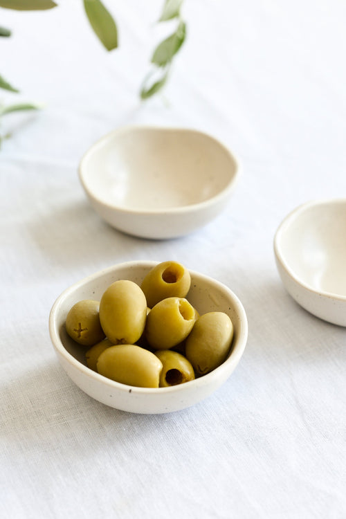 Three small off-white ceramic bowls arranged on a white fabric surface, with one bowl in the foreground holding green olives and two additional bowls blurred in the background. Olive branches appear in the upper left corner.