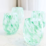 Four stemless wine glasses with a light green and white marbled pattern, arranged on a white marble surface with a soft-focus background.