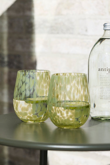 A clear glass bottle labeled 'antipodes' with two textured green drinking glasses on a round dark green side table, set against a light stone wall with a green chair partially visible on the left.
