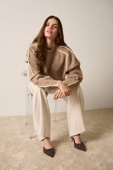 Woman sitting on a chair wearing a brown sweater and beige pants against a plain background