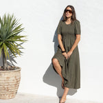 A woman wearing an olive green midi dress and sunglasses stands next to a swimming pool in front of a white wall.