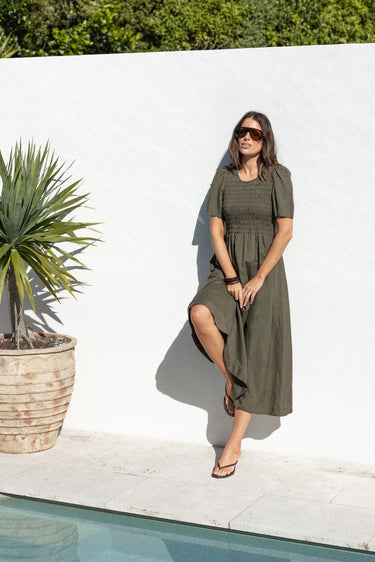 A woman wearing an olive green midi dress and sunglasses stands next to a swimming pool in front of a white wall.