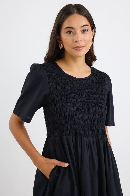 Woman wearing a black midi dress with shirred bodice and short puff sleeves, styled with black sneakers and gold earrings.