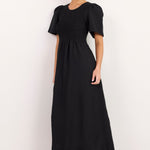 A woman modeling a black midi dress with short puff sleeves, a smocked bodice, and a gathered skirt, paired with black strappy flat sandals against a plain white studio background.