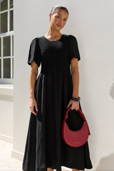 A woman wearing a black midi dress with puff sleeves and a smocked bodice, holding a red crescent-shaped handbag, standing against a white stucco wall.