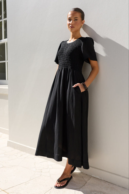 A woman wearing a black midi dress with a smocked bodice and short puff sleeves, leaning against a white stucco wall outdoors.