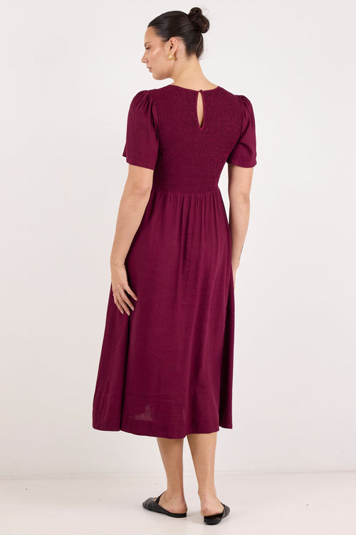 A woman modeling a short-sleeve midi dress in deep burgundy from the back, standing against a plain white studio background.