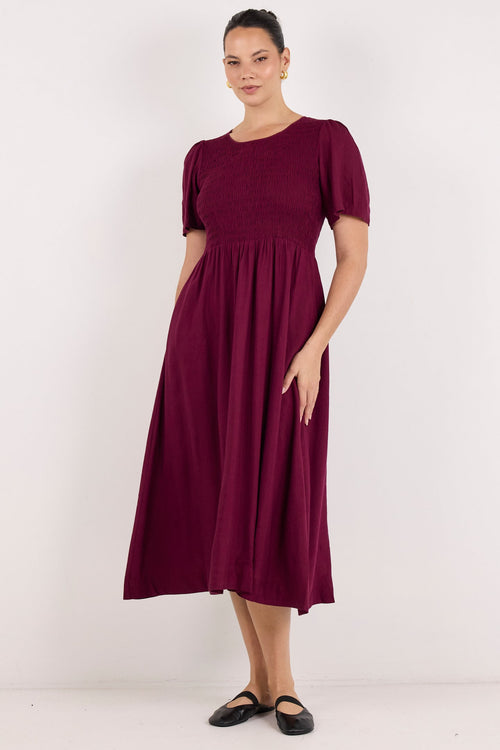 A woman modeling a short-sleeve midi dress in deep burgundy, standing against a plain white studio background.