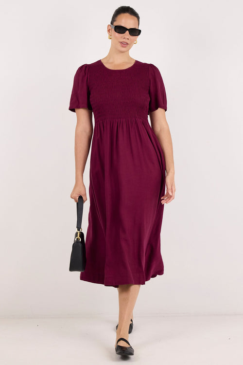 A woman modeling a short-sleeve midi dress in deep burgundy, paired with black sunglasses, black shoes, and a small black handbag, set against a plain white studio background.