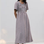 A woman wearing a short-sleeve gingham midi dress and sunglasses stands on a tiled pool deck against a white wall.