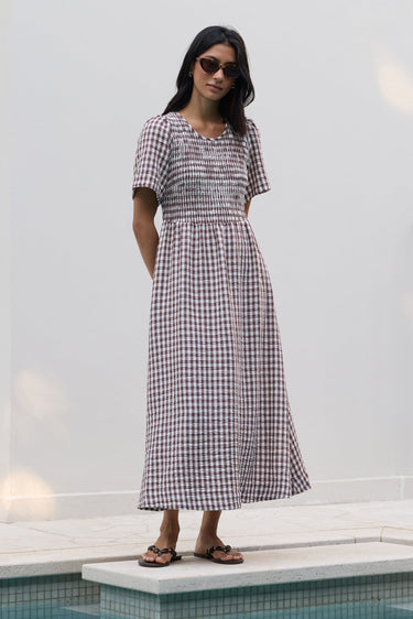 A woman wearing a short-sleeve gingham midi dress and sunglasses stands on a tiled pool deck against a white wall.